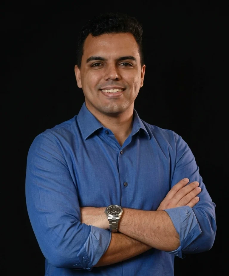 Foto de perfil de Davi Teixeira sorrindo, vestindo camisa social azul e relógio prata, com braços cruzados sobre fundo preto.